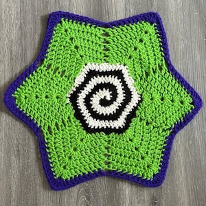 May include: A six-pointed star-shaped crochet piece featuring a vibrant lime green center with a central spiral pattern in black and white yarn. The outer edge of the star is finished with a deep purple border, creating a distinct contrast.