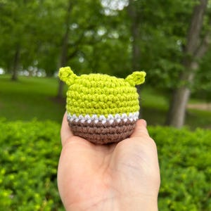 May include: A small, crocheted figure in the shape of a character's head, featuring a light green top, a white and brown midsection, and two small, upright ears. The figure is held in a hand against a blurred green background.