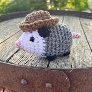May include: A crocheted opossum figurine wearing a brown cowboy hat. The opossum has a gray and white body, black eyes, a pink nose, and a pink tail. The figurine is sitting on a wooden surface.