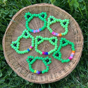 May include: Six green beaded bracelets shaped like frogs, each with black bead eyes and a rainbow-colored section. The bracelets are arranged in a woven basket on a bed of green grass.
