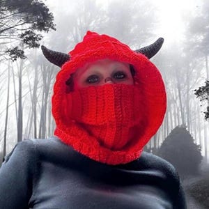 May include: A red crocheted hooded cowl with black horns. The cowl covers the face and neck, with only the eyes visible.