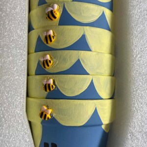 May include: A stack of blue ceramic pots featuring hand-painted yellow scalloped patterns and small three-dimensional bee figures attached to the rims. The bottom pot displays the word Hunn in bold black lettering, all nestled in white foam.