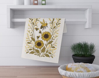 Op de afbeelding: Een witte keukendoek met een zonnebloem- en bijenontwerp. De handdoek heeft twee zonnebloemen, drie bijen en gele en bruine bladeren. De handdoek hangt aan een witte plank boven een houten aanrecht.