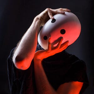 Op de afbeelding: Een wit masker met drie zwarte, cirkelvormige ooggaten wordt door een persoon omhoog gehouden. De persoon draagt een zwart kledingstuk. De afbeelding is verlicht met rood licht, wat een dramatisch effect creëert tegen de donkere achtergrond.