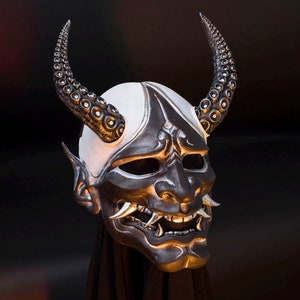 May include: A black and white Oni mask with detailed horns and a fierce expression. The mask is made of a hard material and has a textured surface.