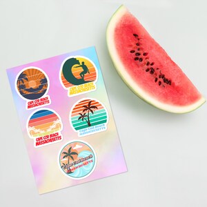 Könnte beinhalten: Fünf runde Aufkleber mit einem Retro-Strandthema. Jeder Aufkleber zeigt ein anderes Design mit einem Sonnenuntergang, Palmen, einem Surfer und Wellen. Die Aufkleber sind mit dem Text "Cape Cod Beach Massachusetts" bedruckt.