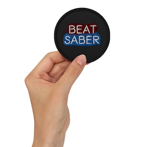 May include: A black round patch with white embroidered text that reads "BEAT SABER". The text is in two lines, with "BEAT" in red and "SABER" in blue.