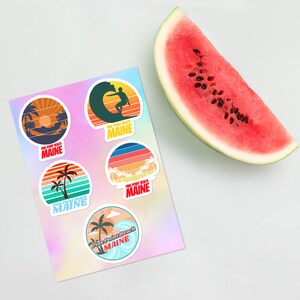 Könnte beinhalten: Fünf bunte runde Aufkleber mit einem Retro-Strandthema. Jeder Aufkleber zeigt eine andere Strandszene mit Palmen, einem Sonnenuntergang und einem Surfer, der auf einer Welle reitet. Der Text "Pine Point Beach Maine" befindet sich auf jedem Aufkleber.