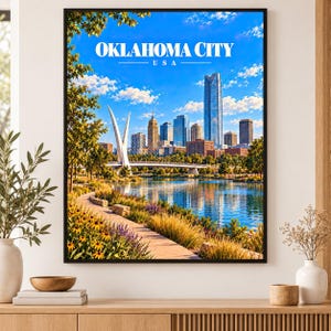 May include: A framed wall art print featuring the Oklahoma City skyline under a bright blue sky with white clouds. The scene includes a river, a white suspension bridge, and a paved path lined with yellow flowers and green trees. Text reads OKLAHOMA CITY USA.