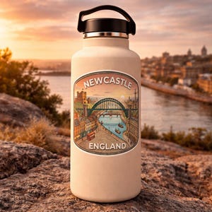 Op de afbeelding: Een beige waterfles met een zwarte handgreep en deksel. De fles heeft een kleurrijke sticker met de tekst "Newcastle England" en een illustratie van een brug, gebouwen en een rivier. De achtergrond toont een zonsondergang boven een stad.
