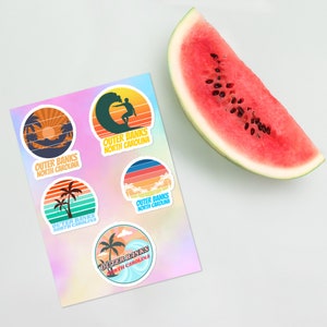 May include: Five colorful round stickers with the text "Outer Banks North Carolina" and various beach-themed illustrations, including palm trees, waves, and a surfer.