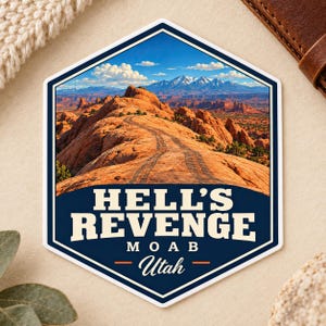Peut inclure: Un autocollant hexagonal représentant un paysage de formations rocheuses rouges et un sentier de terre sous un ciel bleu avec des nuages blancs. Le texte HELL'S REVENGE MOAB Utah est écrit en lettres blanches et crème sur fond bleu.