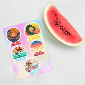 Könnte beinhalten: Fünf bunte runde Aufkleber mit Retro-Design, die Palmen, einen Surfer und den Text "Long Beach California" zeigen.