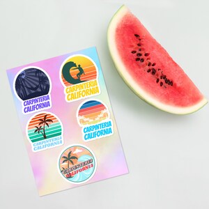 May include: Five colorful stickers with the text "Carpinteria California" and various beach-themed illustrations, including a surfer, palm trees, and waves. The stickers are on a holographic background and a slice of watermelon is in the background.