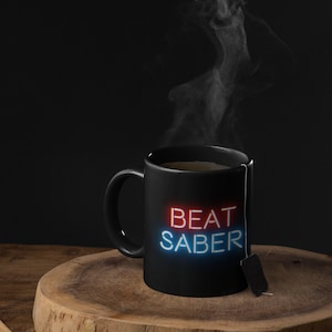 May include: A black mug with the words "BEAT SABER" in red and blue neon lettering. The mug is filled with a steaming beverage and a tea bag hangs from the rim.