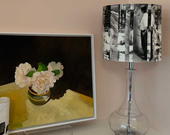 May include: A glass table lamp with a clear glass base and a cylindrical lampshade. The lampshade is covered in a black and white patterned fabric featuring a photo of people walking down a street. The lamp is sitting on a white surface next to a framed painting of white flowers in a green vase.