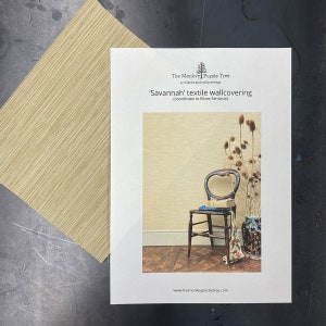 May include: A sample of 'Savannah' textile wallcovering, with a light beige textured surface, next to a larger sample. The image also shows a decorative scene with a chair, vase, and dried plants. The text on the sample reads: 'Savannah' textile wallcovering.