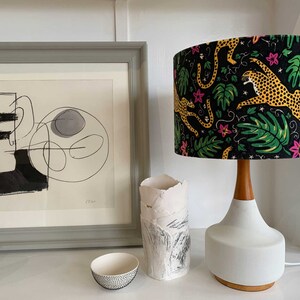 May include: A white ceramic table lamp with a wooden base and a black lampshade featuring a colourful print of leopards and tropical leaves.
