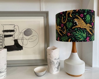 Könnte beinhalten: Eine weiße Keramik-Tischlampe mit Holzsockel und einem schwarzen Lampenschirm mit einem bunten Druck von Leoparden und tropischen Blättern.