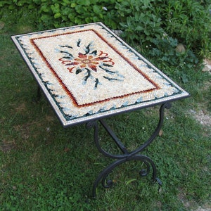 Puede incluir: Una mesa con una tapa de mosaico con un diseño floral en tonos rojos, azules y blancos. La mesa tiene una base de hierro forjado negro.