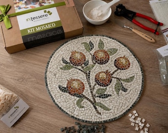 May include: A circular mosaic art piece featuring a pomegranate tree design with red and orange fruit and green leaves on a white background. The kit includes a cardboard box labeled intessere, a white bowl with powder, pliers, and loose tiles.