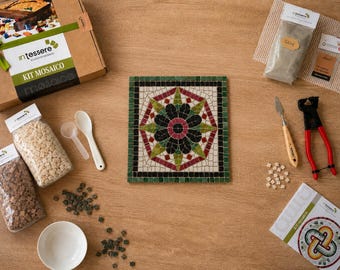 May include: A mosaic craft kit featuring a square tile with a central floral pattern in red, green, and black, surrounded by a green border. The kit includes a cardboard box, bags of small square tiles, a white bowl, a spatula, nippers, and glue.