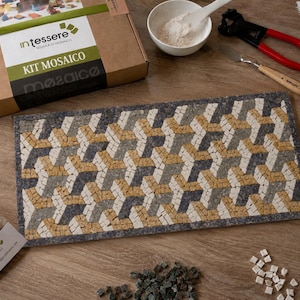 Può includere: Un progetto a mosaico rettangolare con un motivo geometrico di cubi intrecciati nei toni del blu scuro, grigio chiaro e beige. Il kit include una scatola di cartone con l'etichetta intessere KIT MOSAICO, una ciotola bianca con polvere, pinze e una spatola.