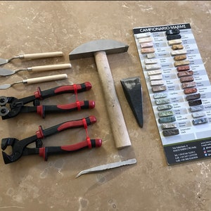 Può includere: Un set di strumenti per lavorare la pietra, tra cui un martello, uno scalpello, delle pinze e una piccola spatola. Gli strumenti sono disposti su una superficie marrone chiaro, insieme a una cartella colori di diversi tipi di marmo.
