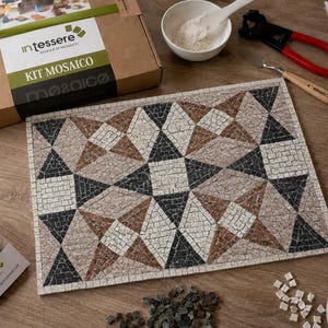 May include: A mosaic craft kit featuring a rectangular board with a geometric pattern in white, black, and brown stone tiles. The kit includes a cardboard box labeled Intessere Kit Mosaico, a bowl of powder, metal nippers, a spatula, and loose tiles.