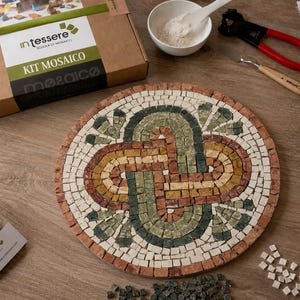 May include: A circular mosaic project featuring an interlocking knot pattern in shades of green, yellow, and terracotta, surrounded by a white border. The kit includes a cardboard box labeled 'intessere KIT MOSAICO', tools, and bags of tiles.