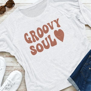May include: A white long-sleeved sweatshirt with a brown graphic that says "Groovy Soul" with a heart next to it. The sweatshirt is laid out on a wooden surface with a pair of jeans and white sneakers.