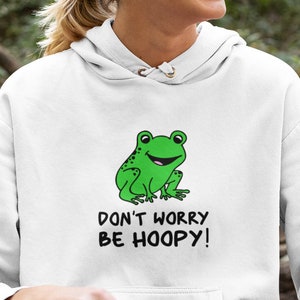 May include: A white hoodie with a green frog illustration and the text "Don't worry be hoopy!" The model is wearing the hoodie and sitting on a log in a forest setting.
