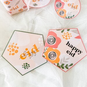 May include: Two pink and white hexagonal cards with green and yellow floral designs. The text "happy eid" is written on each card.