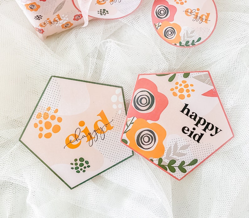 May include: Two pink and white hexagonal cards with green and yellow floral designs. The text "happy eid" is written on each card.