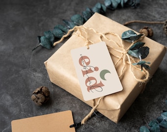 May include: A brown gift box tied with twine and decorated with eucalyptus leaves. A white gift tag with the text "Eid Mubarak" in pink and green lettering and a crescent moon graphic is attached to the box.