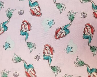 May include: Pink fabric with a repeating pattern of Ariel the Little Mermaid. She is depicted as a mermaid with a green tail and red hair. The pattern also includes starfish and seashells.