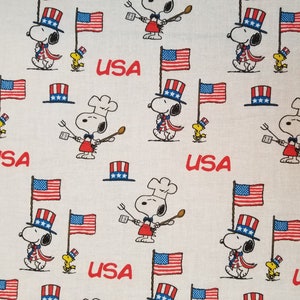 May include: A white fabric with a repeating pattern of cartoon dogs wearing red, white, and blue hats and holding utensils. The dogs are surrounded by American flags and the word "USA" is repeated throughout the fabric.