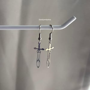 Puede incluir: Un par de pendientes plateados con forma de espada colgados de una barra blanca. Los pendientes tienen un diseño detallado con una cruz en la parte superior y una hoja puntiaguda. El texto "earsbyemilyshop" es visible en la esquina superior derecha.