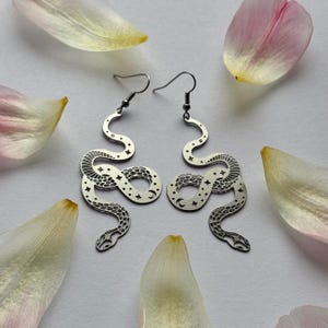 May include: A pair of silver snake earrings. Each earring has a stylized snake design with star and moon cutouts. The earrings are displayed on a white surface, surrounded by pink and yellow flower petals.