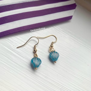 May include: Gold-coloured earrings featuring blue seashell-shaped beads with gold stripes. The earrings are presented on a white surface, with a purple and white striped background. The text "Earsbyemilyshop" is visible.