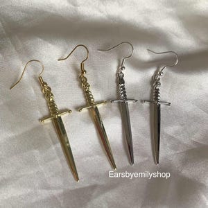 May include: Three pairs of sword-shaped earrings. One pair is gold, one is silver, and one is a darker silver.
