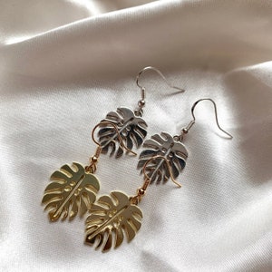 May include: A pair of earrings featuring a gold and silver monstera leaf design. The earrings are made of metal and have a hook fastening.