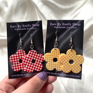 May include: A pair of red and white checked and yellow and white checked flower-shaped earrings with silver hooks. The earrings are displayed on a black card with the text "Ears By Emily Shop Handmade Jewellery" and a website address.
