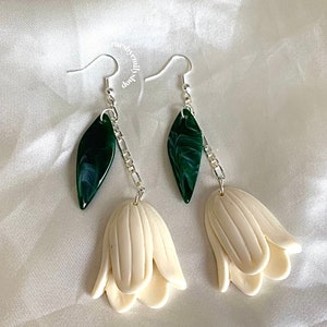 Może przedstawiać: Para białych kolczyków w kształcie kwiatów z zielonymi, marmurowymi liśćmi wiszącymi na srebrnych łańcuszkach. Kolczyki mają delikatny i kobiecy design.