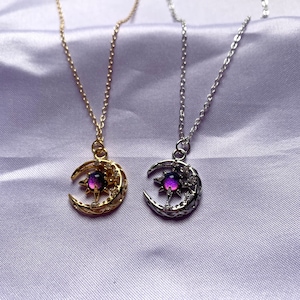Op de afbeelding: Twee kettingen met hemelse thema's. Eén heeft een goudkleurige ketting en hanger met een halve maan en ster ontwerp, met een paarse en roze steen. De andere ketting heeft een zilverkleurige ketting en hanger met een vergelijkbaar ontwerp.