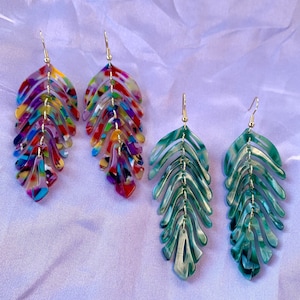 May include: A pair of colourful, multi-layered leaf-shaped earrings and a pair of green, multi-layered leaf-shaped earrings. The earrings are made of acrylic and have a silver tone.