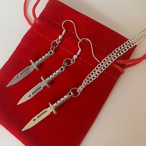 May include: A silver necklace and earrings set featuring a dagger design. The necklace and earrings are displayed on a red velvet pouch.