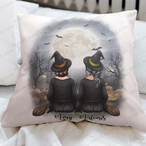 May include: A white pillow with a Halloween-themed design featuring two witches with black hats and broomsticks. The witches are standing in front of a full moon and pumpkins. The text "Amy Artemis" is printed below the witches.