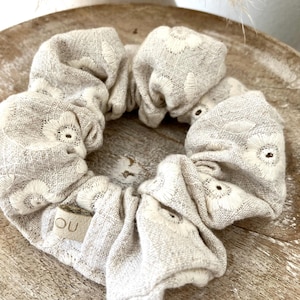 Op de afbeelding: Een beige scrunchie met witte bloemenborduurwerk. De scrunchie is gemaakt van een textuurstof en heeft een klein rechthoekig label met de letters "OU". De scrunchie is een haaraccessoire.