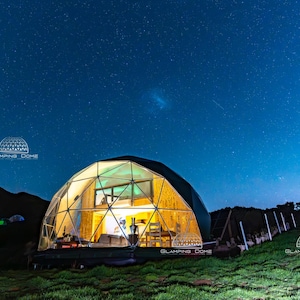Könnte beinhalten: Ein von innen beleuchteter Glamping-Dom mit geodätischem Design vor einem dunklen Sternenhimmel. Der Dom hat eine transparente Front, die eine Innenausstattung zeigt. Der Text "GLAMPING DOME" ist sichtbar.